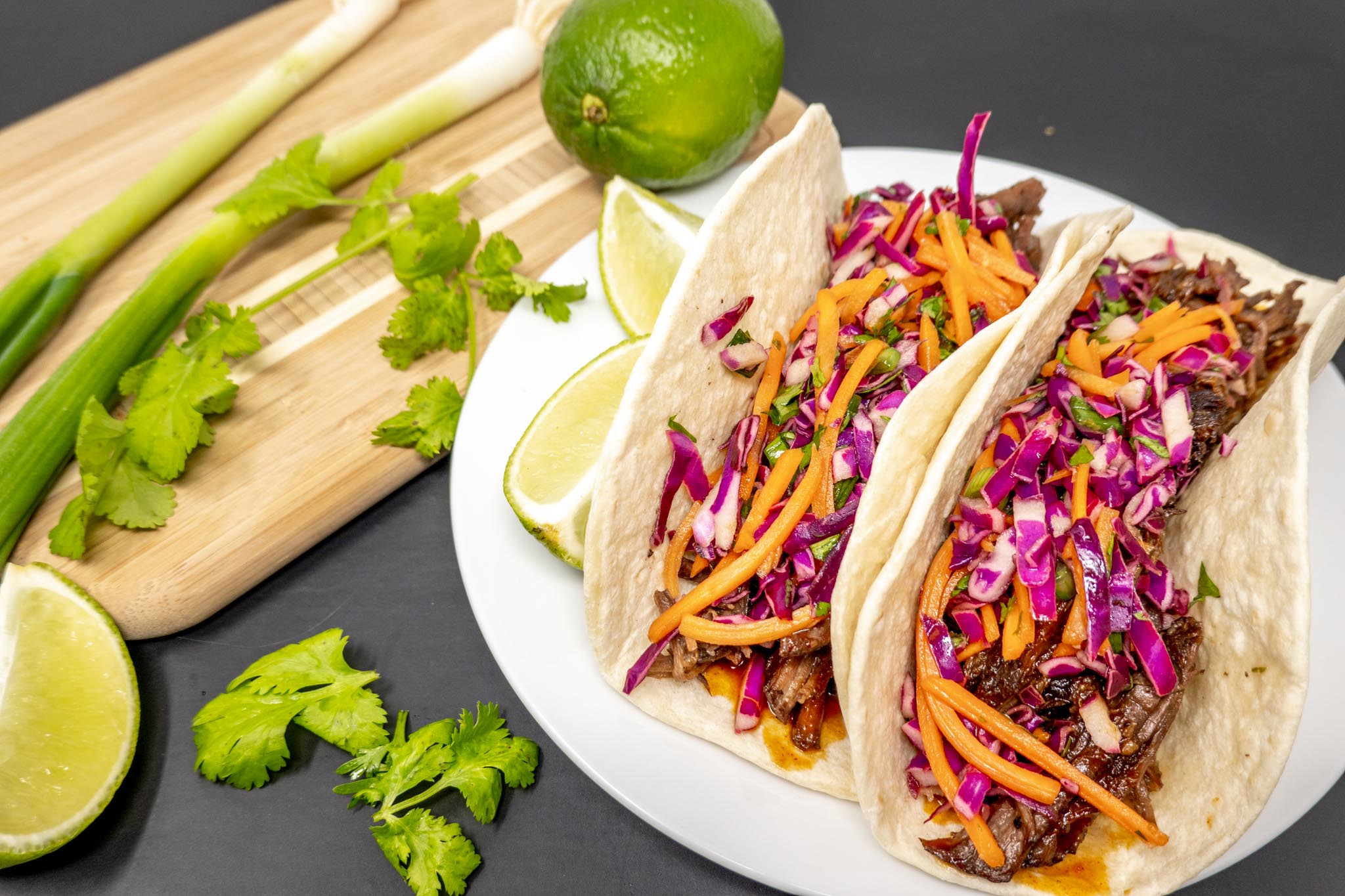 Slow cooker Korean beef tacos on plate next to limes, cilantro, and scallions