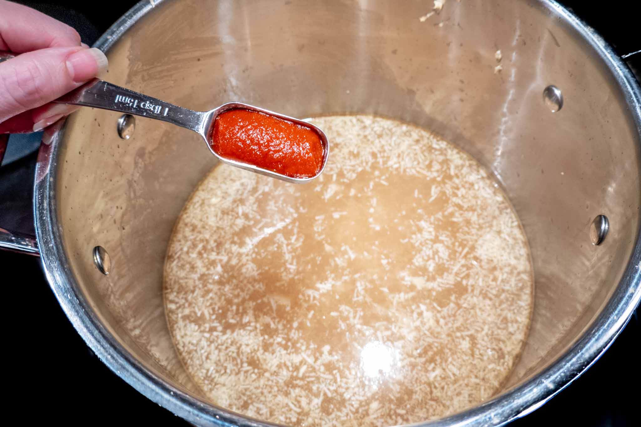 Person holding a tablespoon of siracha over a pot of broth
