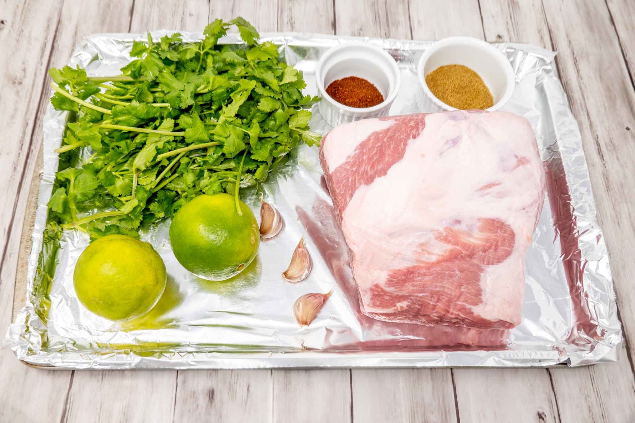 Pork shoulder, spices, garlic, limes, and cilantro on a foil-covered tray