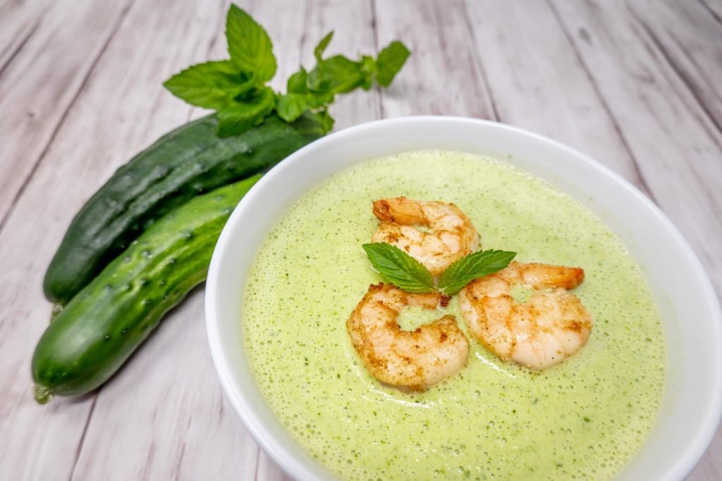 Bowl of green gazpacho topped with shrimp next to cucumbers and herbs