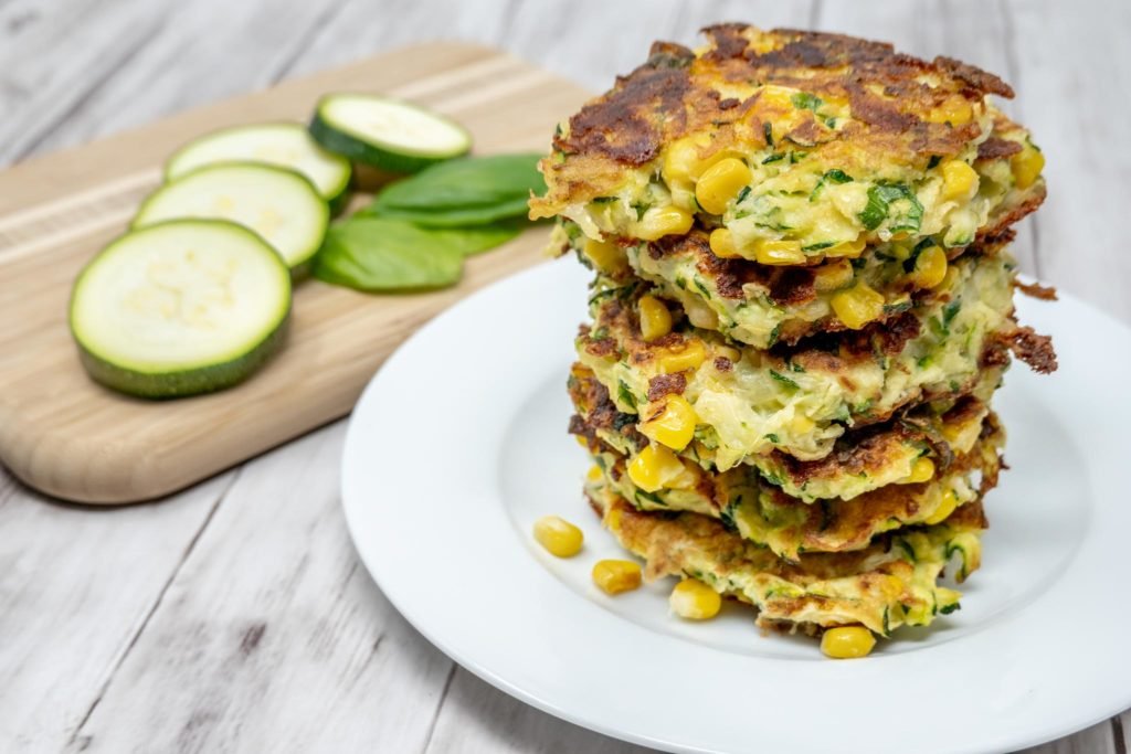 Stack of corn and zucchini fritters on a plate beside slices of zucchini