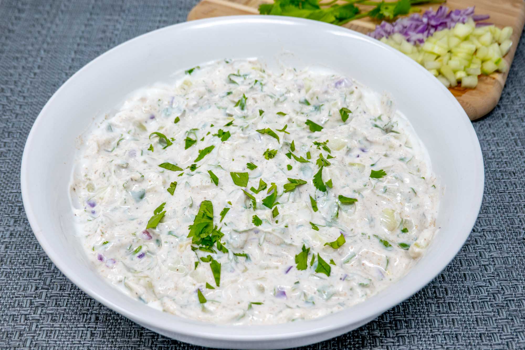 Bowl of yogurt dip beside a cutting board with cucumber, onion, and cilantro