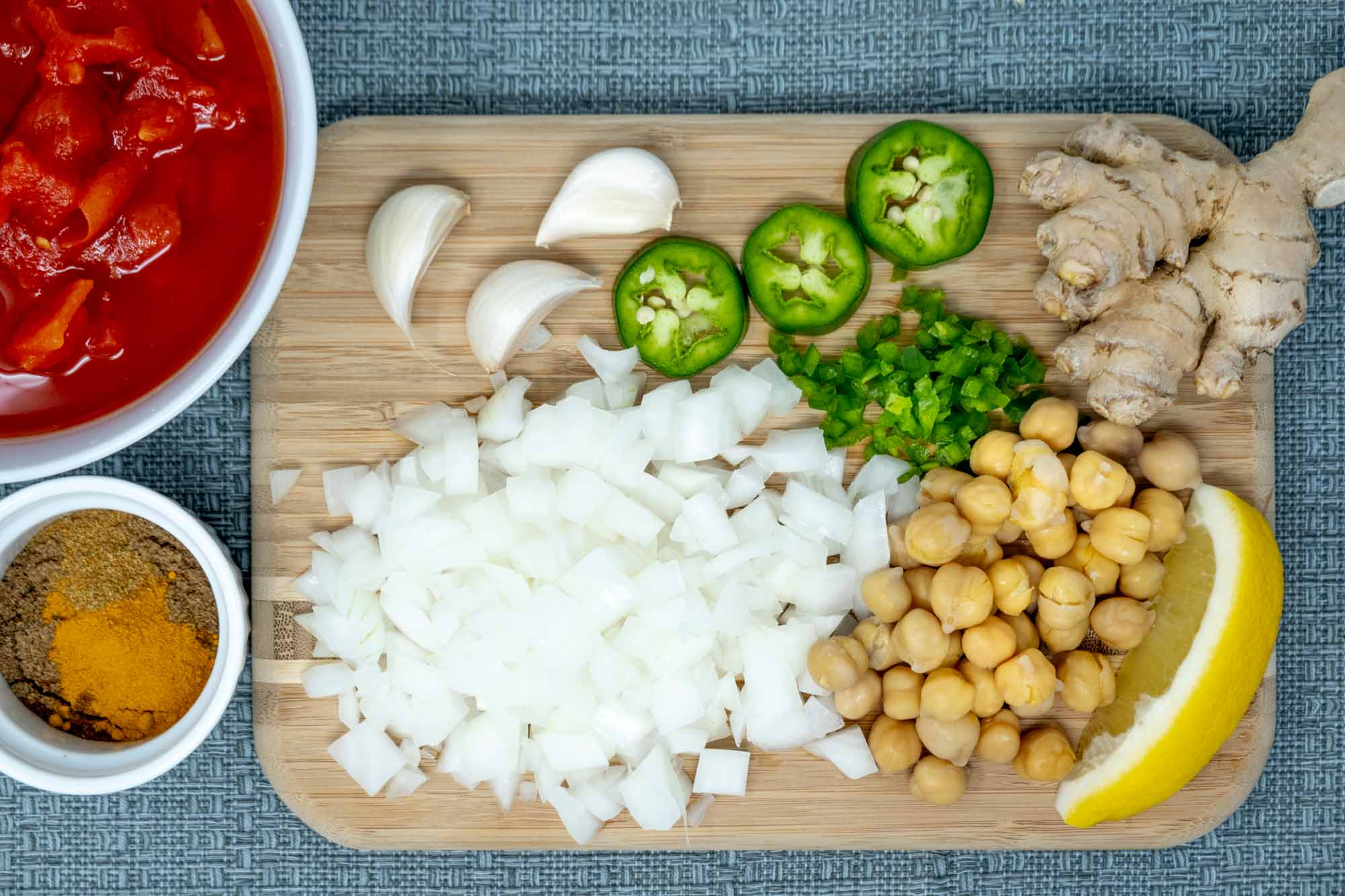 Ginger, garlic cloves, jalapeno, chickpeas, lemon, and diced onion on a cutting board beside bowls of spices and diced tomatoes