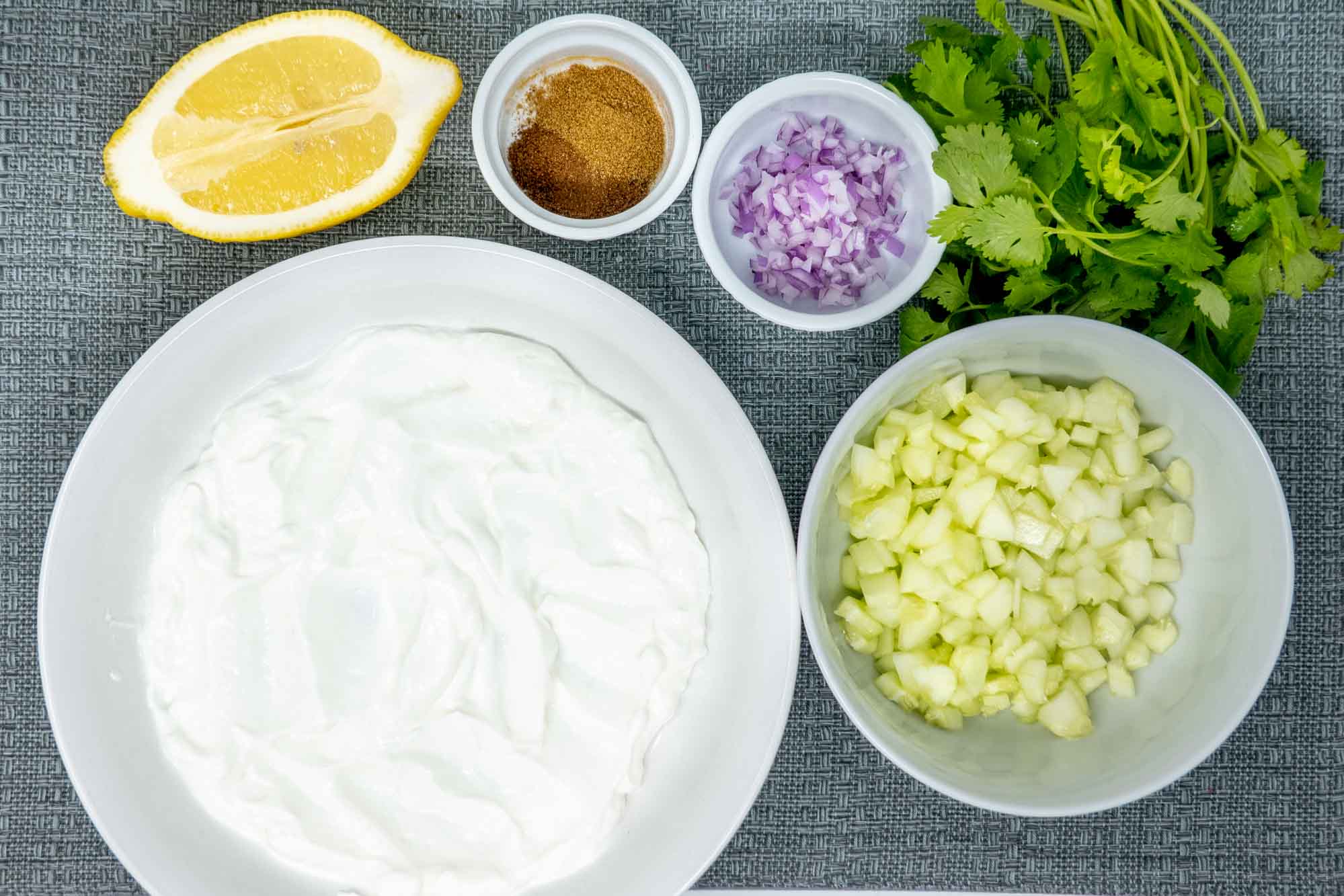 Bowls of Greek yogurt and chopped cucumber on a table next to half a lemon, spices, minced onion, and cilantro
