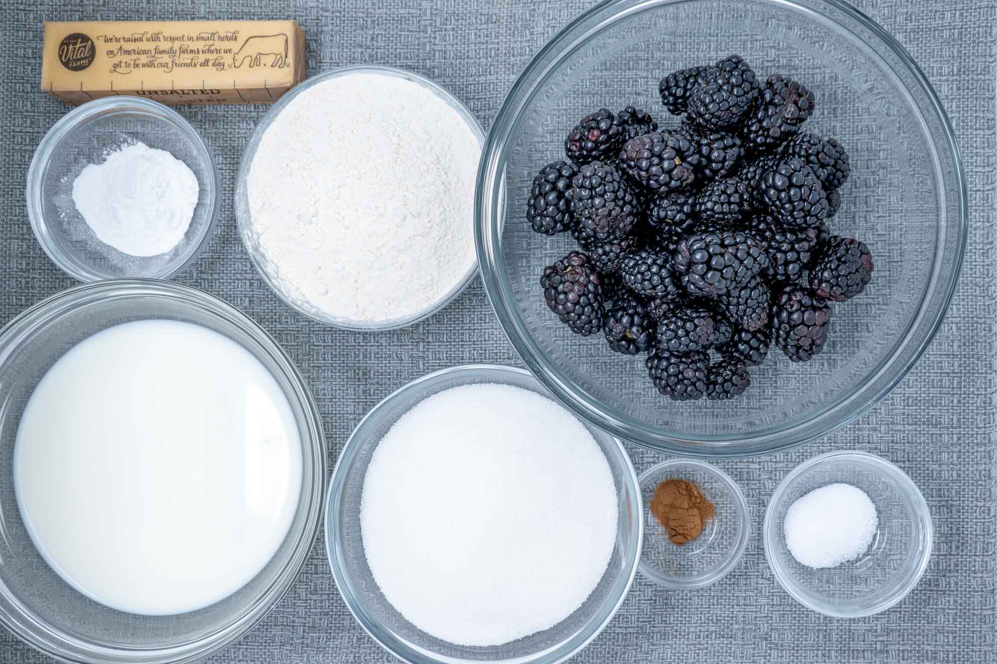 Blackberries, cinnamon, sugar, stick of butter, and other ingredients in bowls on a table