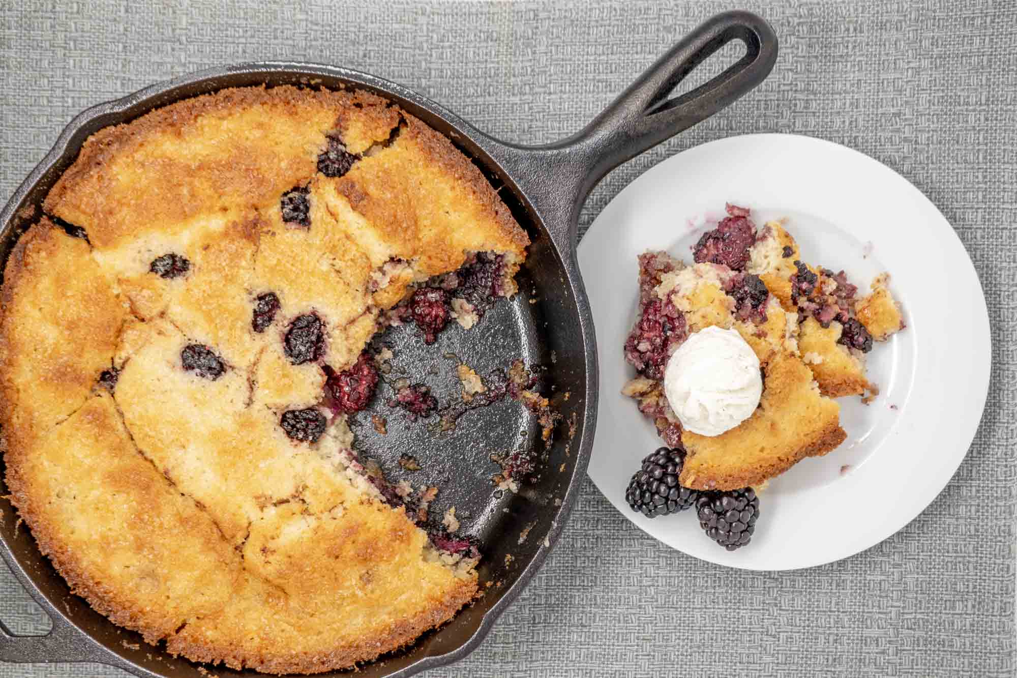 Portion of blackberry cobbler topped with ice cream on a plate next to the rest of the cobbler in a cast iron skillet 