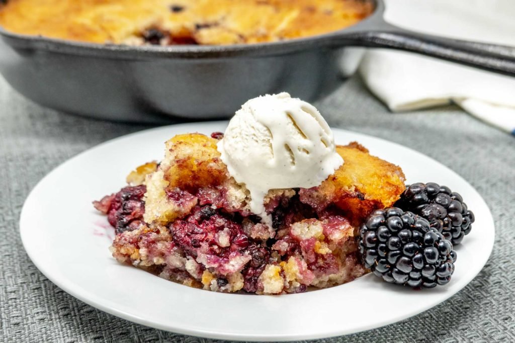 blackberry cobbler topped with melting ice cream on a plate in front of a cast iron skillet with the rest of the cobbler