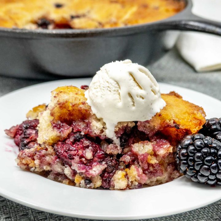 blackberry cobbler topped with melting ice cream on a plate in front of a cast iron skillet with the rest of the cobbler