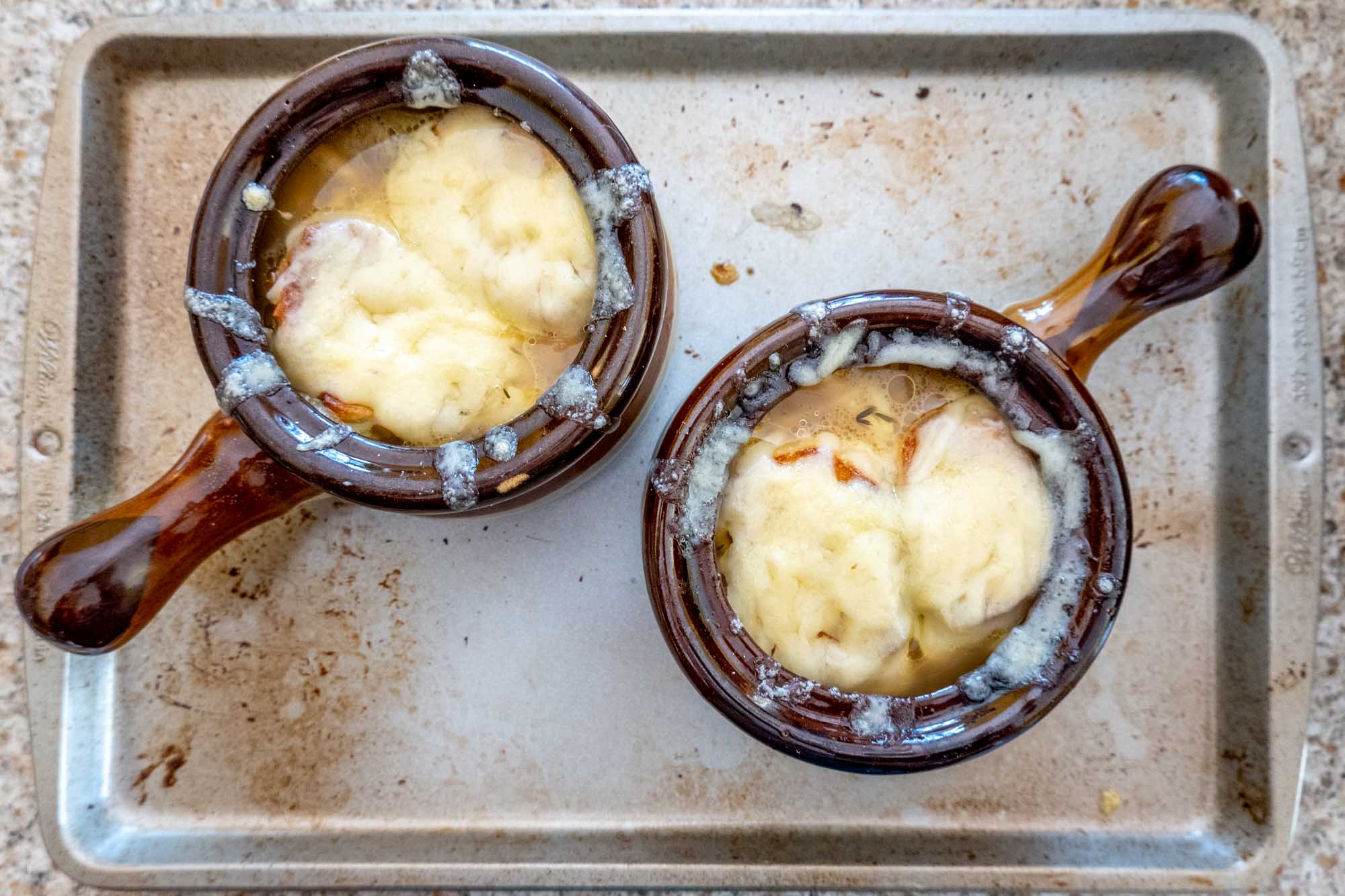 Two soup crocks topped with cheese on a baking sheet