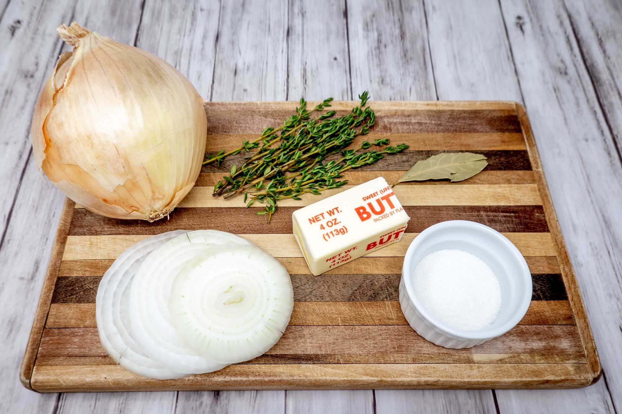 Onions, butter, thyme sprigs, sugar, and a bay leaf on a cutting board