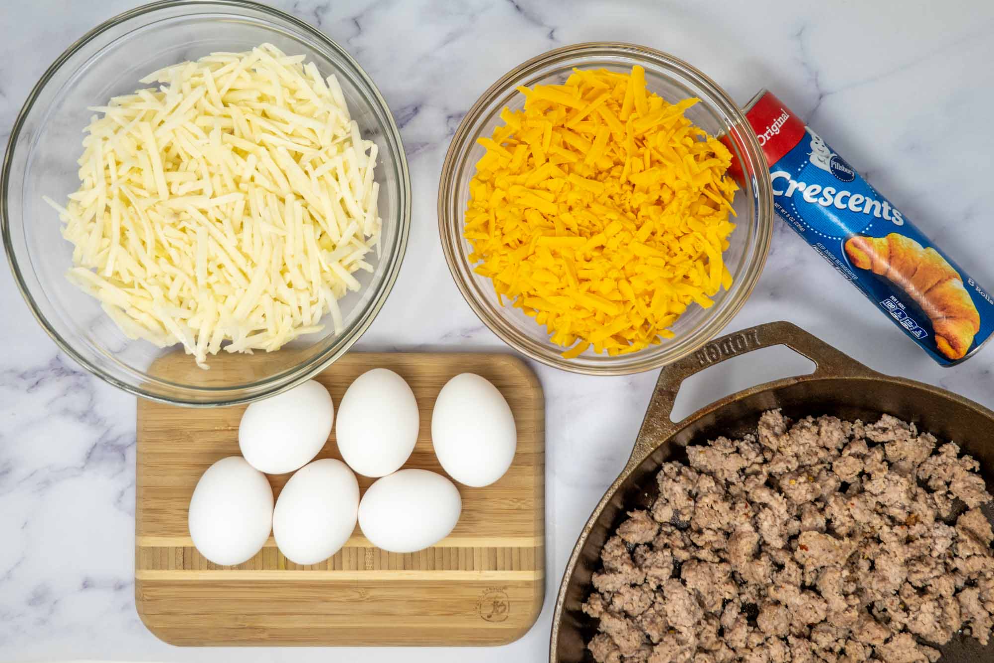 Shredded hash browns, shredded cheese, can of crescent rolls, six eggs, and skillet of cooked sausage.